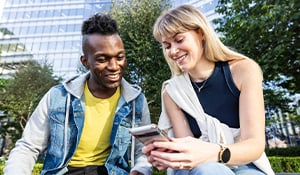 Two young adults sitting outdoors near modern office buildings, smiling and looking at a smartphone together. One wears a yellow shirt and denim jacket, the other a sleeveless top with a white sweater over their shoulders.