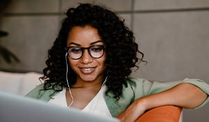A woman with curly hair and glasses is sitting on a couch, smiling while looking at her laptop. She is wearing earphones and appears to be engaged in a virtual meeting or video call in a modern, relaxed indoor setting.