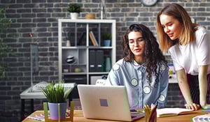 Two women working together at a laptop in a modern office with a brick wall and potted plant.